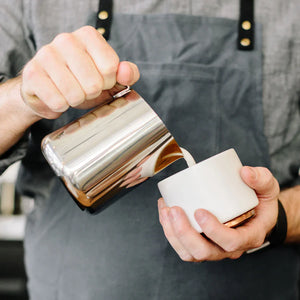 Barista kaataa maitoa kiillotetusta ruostumattomasta teräksestä valmistetusta kannusta, osa Espresso-tarvikkeita ja maitokannua, kaikki kahvista.