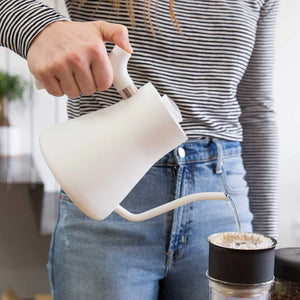 Käsi kaataa vettä valkoisesta kapeakärkisestä vedenkeittimestä kahvinsuodattimeen, osa EspressoGearia, kaikki kahvista.